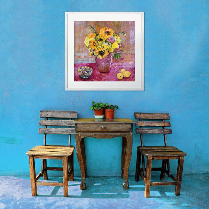 Sunflower prints showing bright sunflowers in a vase on a table with a pink cloth. The sunflower art print is framed on a wall above a table and chairs. The sunflower wall art is available from Judi Glover Art