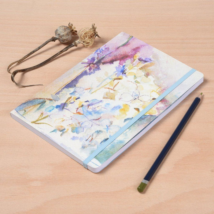 The floral notebook by Judi Glover Art on a table next to a pencil for scale. The notebook is a5 in size and each artistic notebook has 120 lined pages with an elastic enclosure