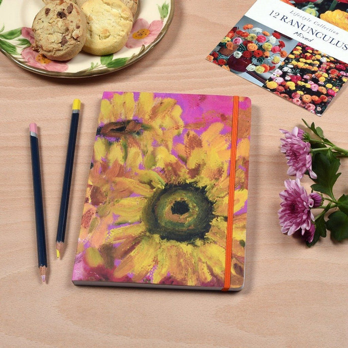 Floral notebook by Judi Glover Art shown on a table surrounded by flowers. The beautiful notebook has sunflowers and a pink background