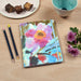 A pretty notebook by Judi Glover Art placed on a table with flowers and pencils. The A5 notebook has a pretty cover with wildflowers and is lined with 120 pages